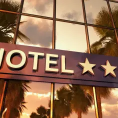 Elegancki hotel z napisem "HOTEL" i czterema gwiazdkami. W szklanej fasadzie odbijają się palmy i zachodzące słońce.