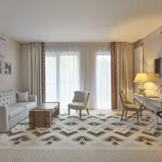 Elegancki pokój w hotelu 5 gwiazdkowym: sofa, stolik, fotele, biurko i TV. Wzory na dywanie i zasłonach dodają charakteru.