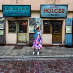 Kobieta w kolorowej sukience pozuje przed sklepami z szyldami "Stanisław Nowak Sklep Spożywczy" i "Benjamin Holcer Stolaz". To jedne z tych fajnych miejsc w Krakowie, które warto odkryć.