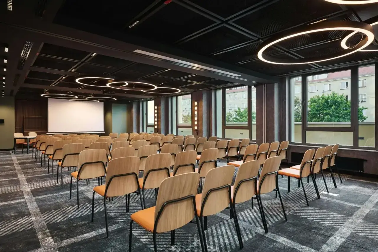 Nowoczesna sala konferencyjna w Sheraton Wrocław. Rzędy drewnianych krzeseł z pomarańczowymi siedziskami, ekran projekcyjny i designerskie oświetlenie.