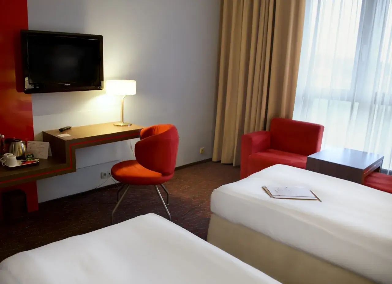Pokój hotelowy 4-gwiazdkowy z dwoma łóżkami, czerwonym fotelem, biurkiem z lampką i telewizorem.