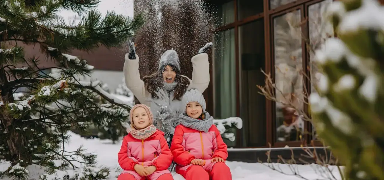 Radosne ferie zimowe! Mama rzuca śnieżkami w dwie uśmiechnięte dziewczynki w różowych kombinezonach, siedzące w śniegu.
