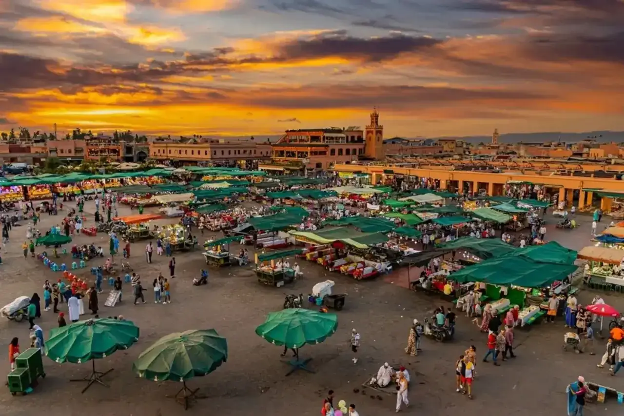 Tętniący życiem plac w Marrakeszu o zachodzie słońca. Ludzie spacerują między straganami pod zielonymi parasolami. Czy w Maroko jest bezpiecznie? Tak, to tętniące życiem miejsce.