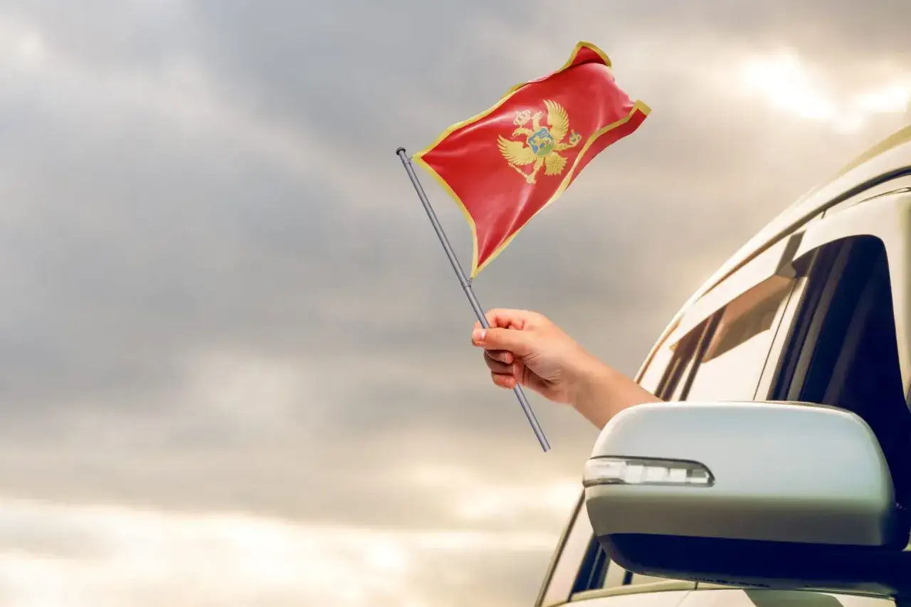 Ręka trzyma flagę Czarnogóry, wyjeżdżając z samochodu. Może to symbolizować podróż lub dumę narodową, jakby to był paszport do wolności.
