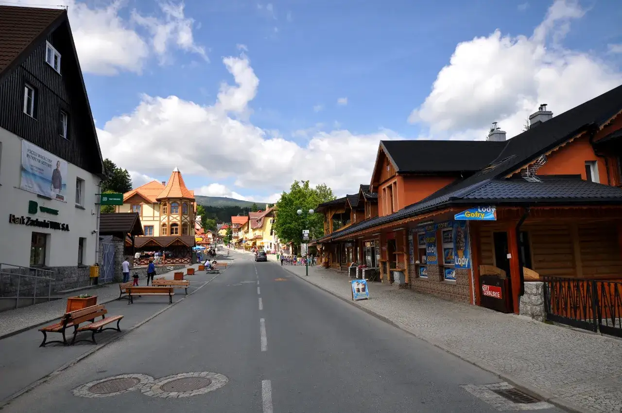 Ulica w Karpaczu centrum, z kolorowymi kamienicami, sklepikami i ławkami. Słoneczny dzień, chmury na niebie.