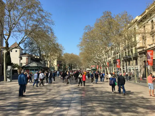 Tłum ludzi spaceruje po słynnej alei **La Rambla w Barcelonie**, otoczonej drzewami i budynkami.